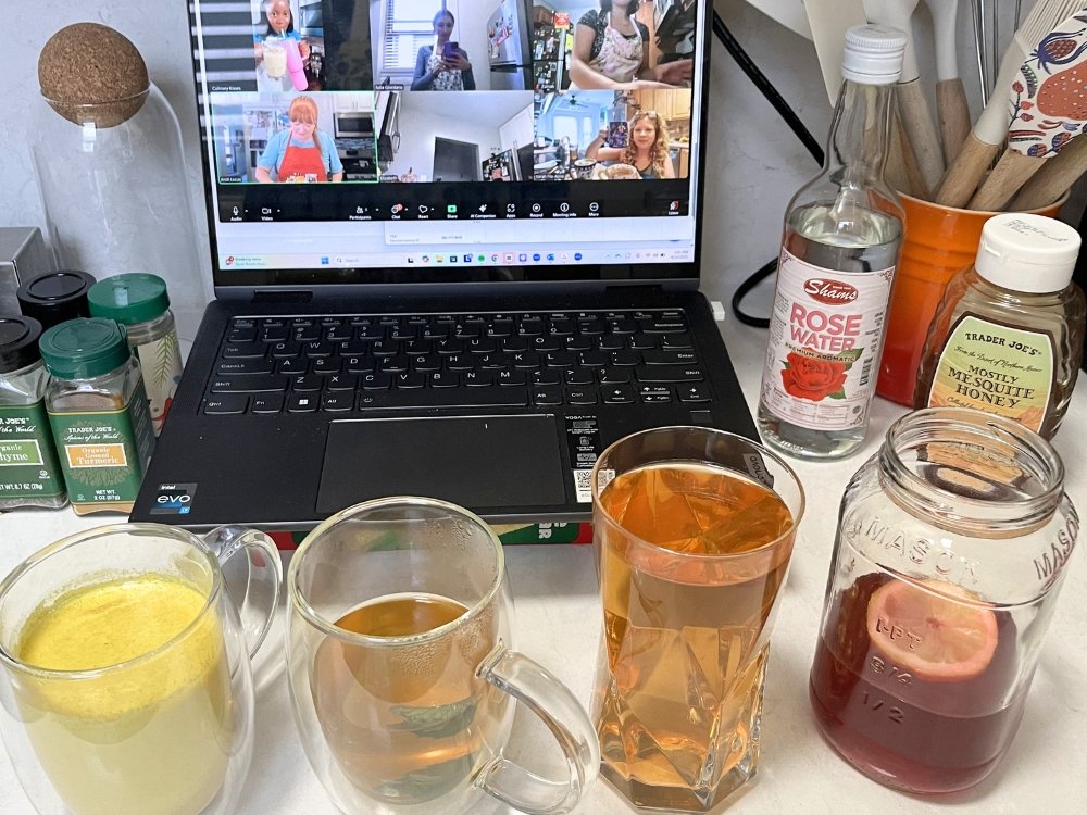 Four glasses with different colored drinks in each sit in front of a laptop that is open to a Zoom meeting with multiple people.
