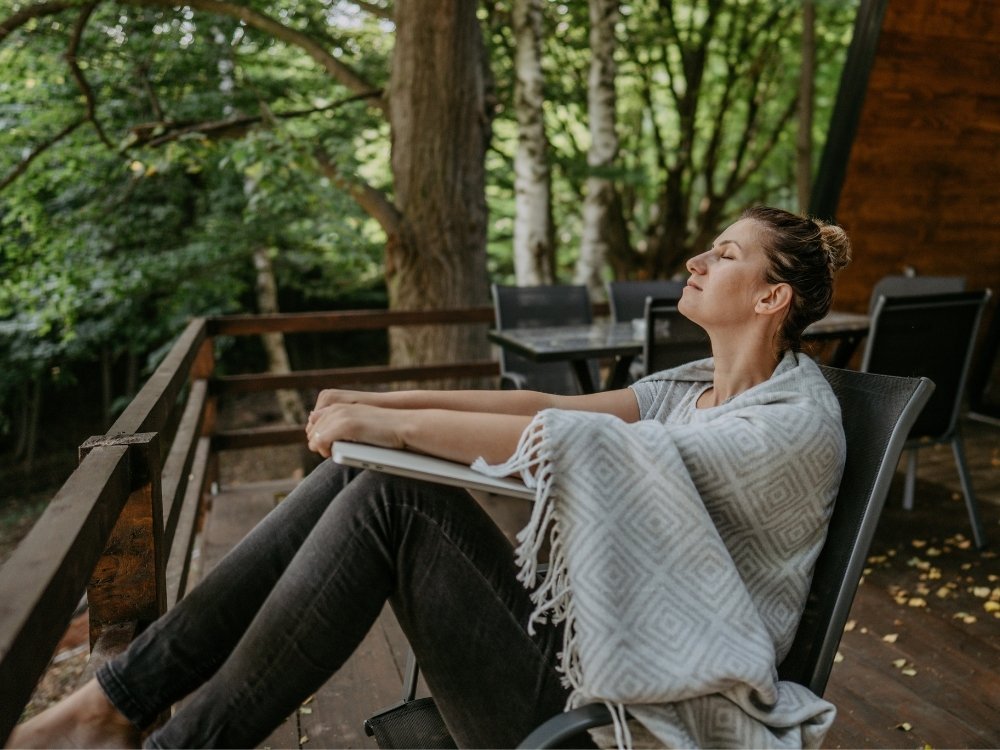 A woman relaxing on a deck surrounded by trees with her laptop shut. The tip to take regular breaks throughout your work day from the blog, How to Avoid Burnout, Writer’s Block, and Creative Slumps by Hummingbird Marketing Services.