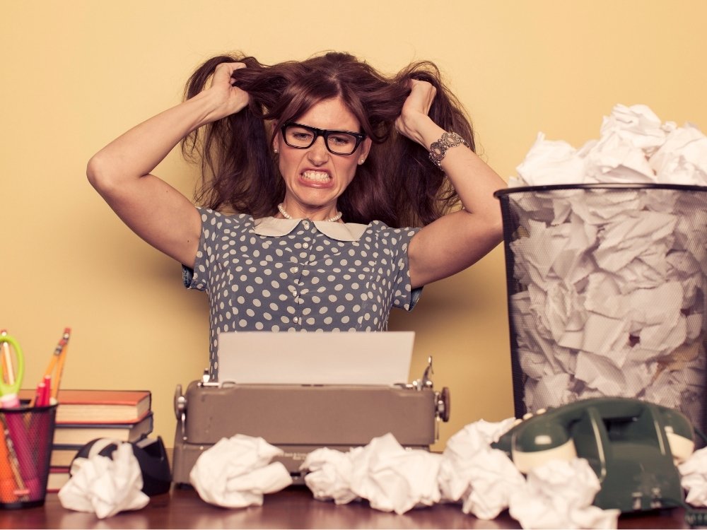 A woman sitting at her desk, frustrated and pulling her hair due to writer’s block, with a stack of crumpled-up paper in the trash can next to her. Avoid feeling this way with tips from the blog, How to Avoid Burnout, Writer’s Block, and Creative Slumps by Hummingbird Marketing Services.