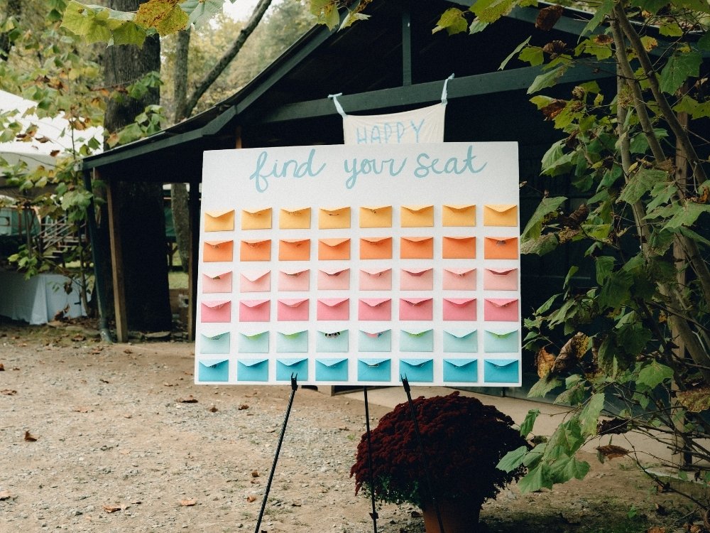 An example of a DIY seating chart idea: A large white sign that says "find your seat" with different colored envelopes arranged in rows of the same color with yellow, orange, pink, and blue envelopes.