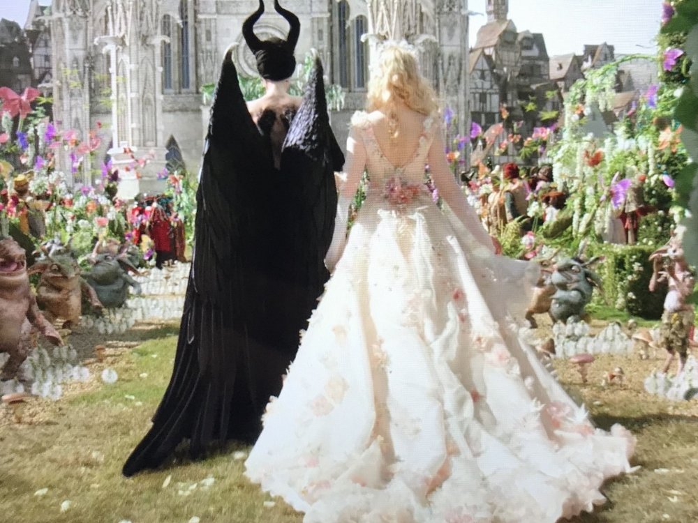 Maleficent in a black gown walking down an aisle with a bride in a white and floral gown towards a fairytale castle.