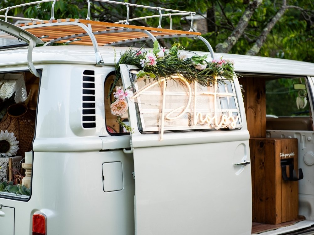 An old van converted to a photo booth for events, with flowers and a neon sign that says "photos" on the outside.