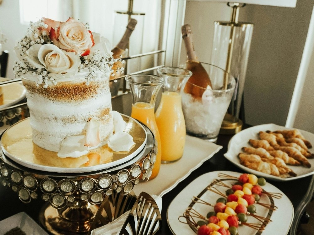 A wedding cake with flowers on top, along with two plates of appetizers, two carafes of orange juice, and bottle of wine on ice.