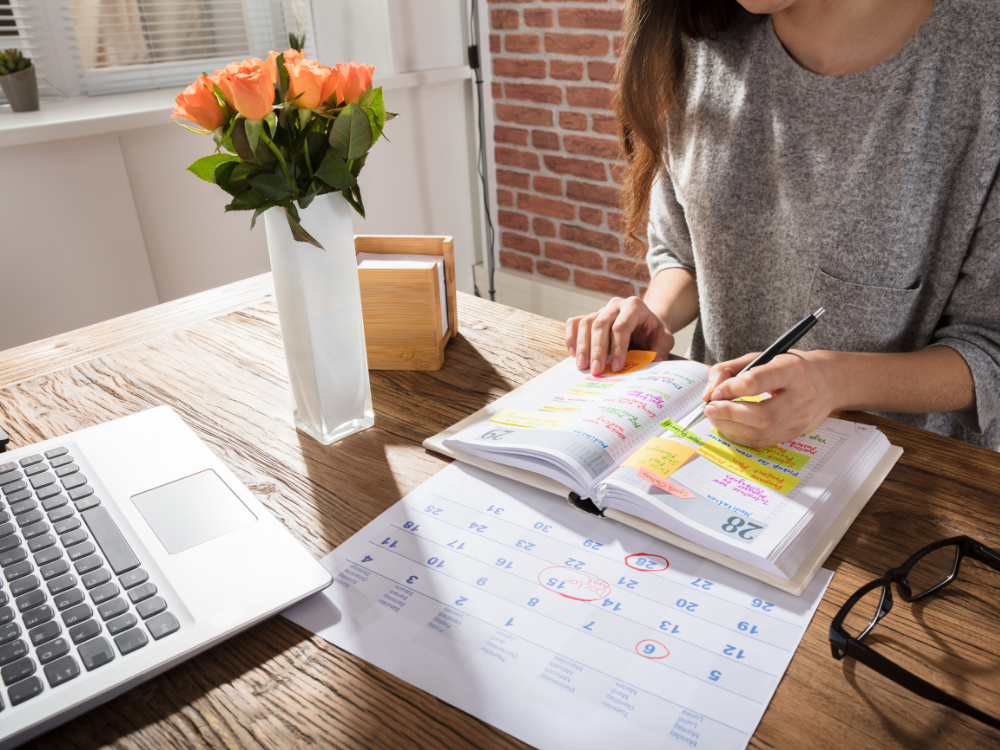 A desk with a laptop, calendar, planner, and vase of flowers. A woman sits at the desk with her pen, writing in the planner.