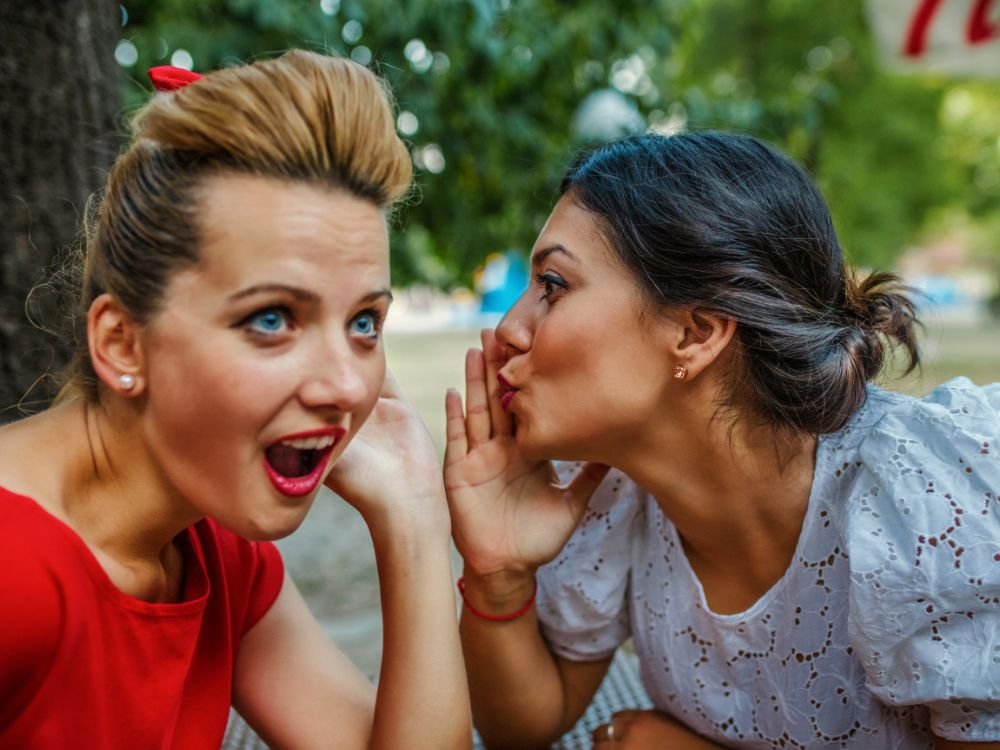 Two women smiling, one woman is whispering something in the other woman's ear.